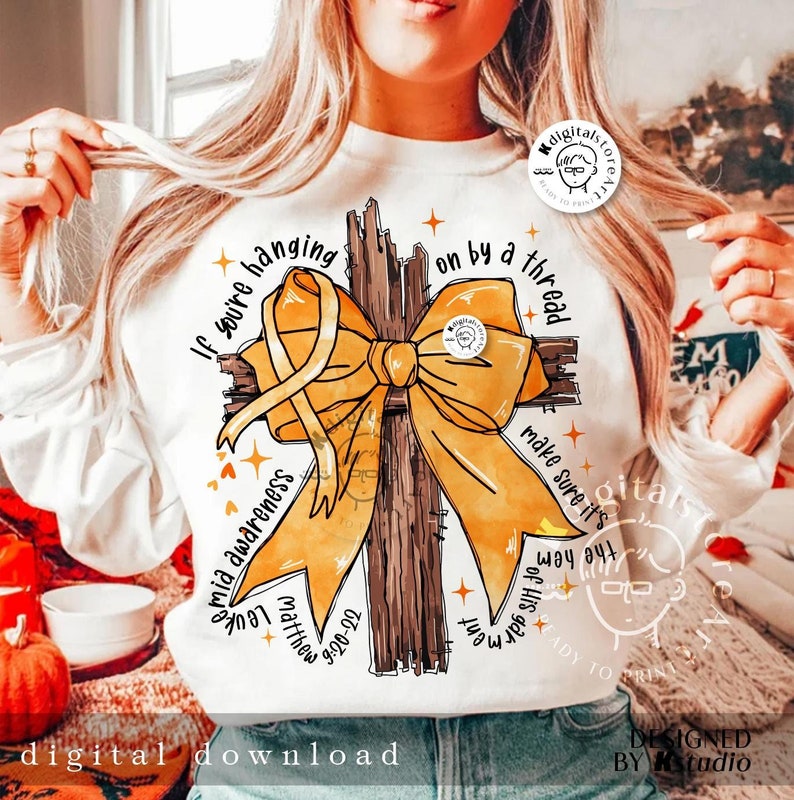 May include: A white sweatshirt with a yellow ribbon tied in a bow around a brown wooden cross. The text "If you're hanging on by a thread make sure it's the hem of His garment" is printed around the cross. The text "Matthew 9:20-22" is printed below the cross.
