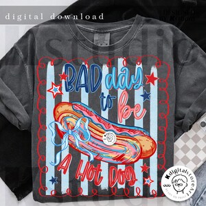 May include: Dark gray t-shirt with a graphic of a hot dog and the text "BAD day to be A Hot Dog". The design includes red, white, and blue colors with stars and stripes, giving it a patriotic feel.