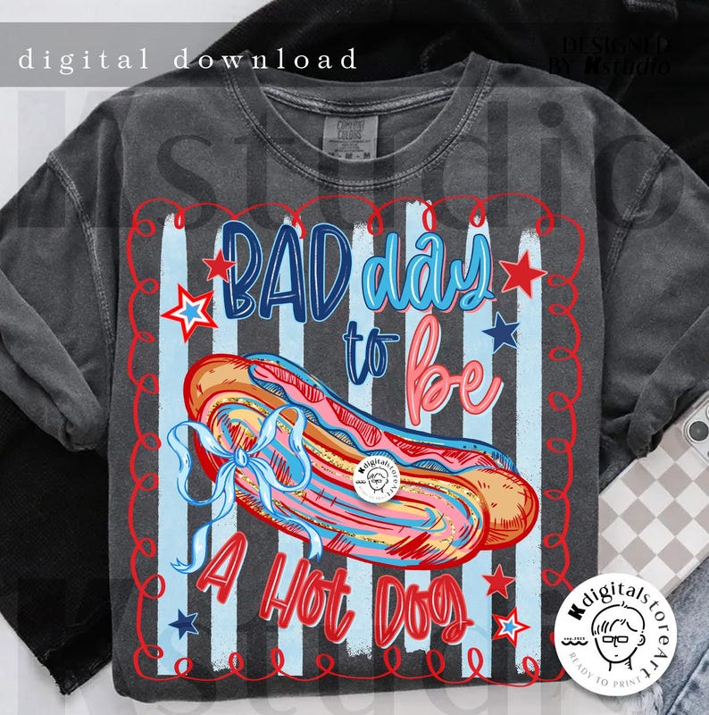 May include: Dark gray t-shirt with a graphic of a hot dog and the text "BAD day to be A Hot Dog". The design includes red, white, and blue colors with stars and stripes, giving it a patriotic feel.