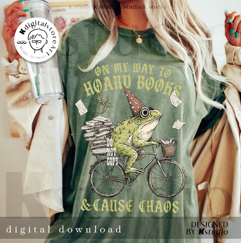 May include: Green t-shirt featuring a frog on a bicycle, wearing a wizard hat, and carrying books. The text reads "ON MY WAY TO HOARD BOOKS & CAUSE CHAOS." The design includes a dragon and flying papers. "Digital Download" is printed at the bottom of the shirt.