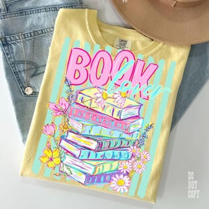 May include: A yellow t-shirt with the words "BOOK LOVER" in pink and blue, with a stack of colorful books and flowers. The shirt is paired with blue jeans and a brown hat.