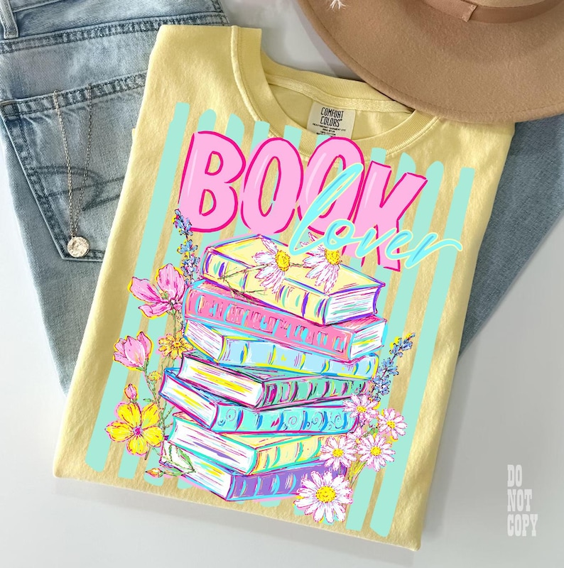 May include: A yellow t-shirt with the words "BOOK LOVER" in pink and blue, with a stack of colorful books and flowers. The shirt is paired with blue jeans and a brown hat.
