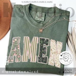 May include: An olive green t-shirt with the word "AMEN" in a floral design. The letters are outlined with a scalloped edge. A gold necklace with a pendant rests on the shirt. The shirt also has the text "digital download" and "DESIGNED BY Kstudio".