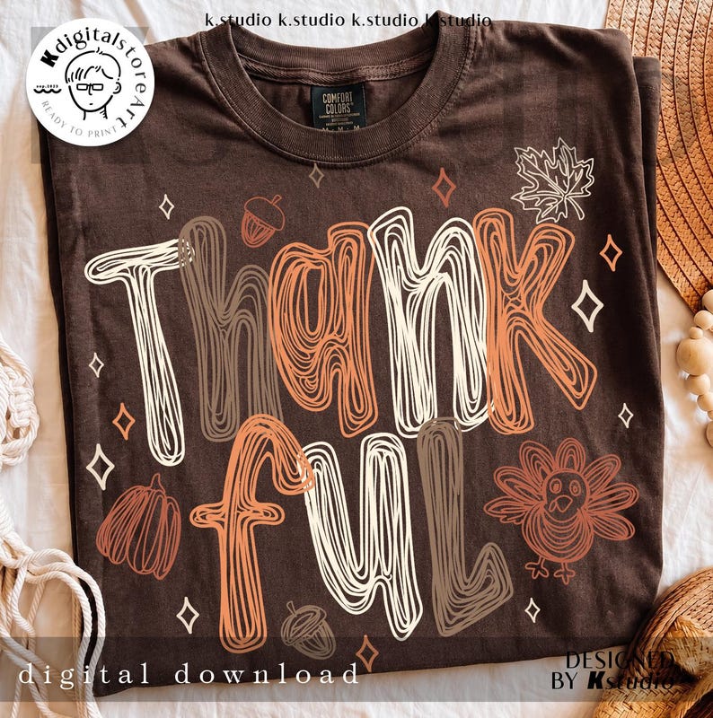 K&ouml;nnte beinhalten: Ein dunkelbraunes Kurzarm-T-Shirt mit dem Wort Thankful in einer gekritzelten Schriftart in Wei&szlig;, Orange und Braun. Das Design enth&auml;lt grafische Illustrationen eines Truthahns, eines K&uuml;rbisses, von Eicheln, Ahornbl&auml;ttern und kleinen Rauten.