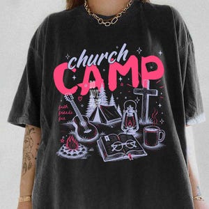 K&ouml;nnte beinhalten: Schwarzes T-Shirt mit einem Grafikdesign mit den Worten "church CAMP" in Pink, zusammen mit Illustrationen einer Gitarre, eines Zeltes, eines Kreuzes, einer Laterne, eines Lagerfeuers, eines Buches und einer Kaffeetasse. Die Worte "faith friends fun" sind ebenfalls enthalten.