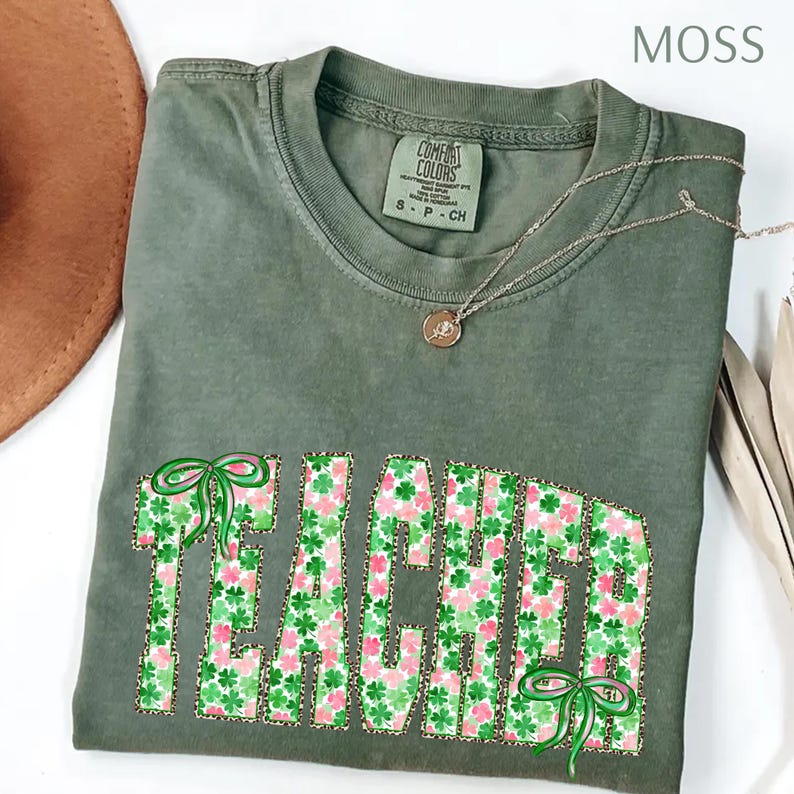 May include: A moss green Comfort Colors t-shirt with the word "TEACHER" in a floral and leopard print design. The shirt features green bows and a delicate gold necklace. The shirt is a small size.