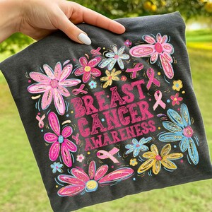 May include: Dark gray t-shirt with a floral design and the words "Breast Cancer Awareness" in pink. The flowers are pink, blue, and yellow, with pink ribbons. A hand is holding the shirt.