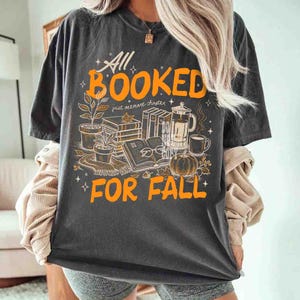 May include: Dark gray t-shirt with an orange graphic that reads "All Booked For Fall." The design includes books, a pumpkin, coffee, and potted plants. The phrase "just one more chapter" is also included.