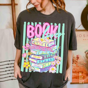 May include: Dark gray t-shirt featuring a colorful graphic of stacked books with flowers and the words "BOOK lover" in pink and light blue. The design is set against vertical mint green stripes.