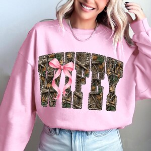 Puede incluir: Sudadera rosa claro con la palabra "WIFEY" en un patr&oacute;n de camuflaje y un lazo rosa. La sudadera es de color rosa claro liso.