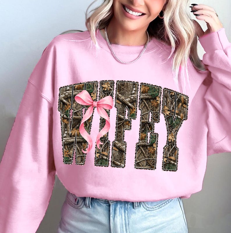 Puede incluir: Sudadera rosa claro con la palabra "WIFEY" en un patr&oacute;n de camuflaje y un lazo rosa. La sudadera es de color rosa claro liso.