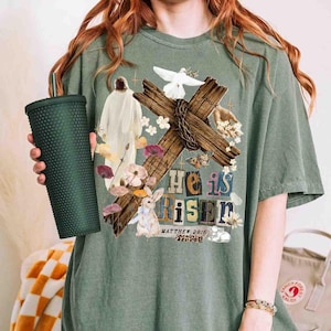 May include: Sage green t-shirt featuring a graphic design with a wooden cross, the words "He is Risen", a dove, and a bunny. The design includes floral and butterfly accents. A dark green textured tumbler is held in the left hand.