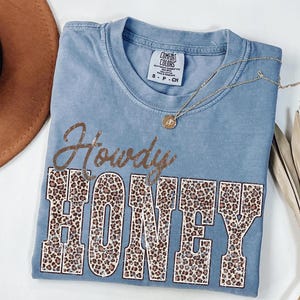 May include: A blue Comfort Colors t-shirt with the words "Howdy HONEY" in a brown, glittery font with leopard print letters. A gold necklace with a small pendant rests on the shirt. The shirt is folded on a white surface.