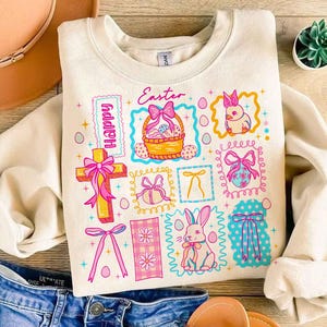 May include: Cream-colored sweatshirt with a colorful Easter-themed graphic. The design includes a cross, a basket of eggs, a bunny, and the word "Easter". The graphic is framed with pink and blue borders.