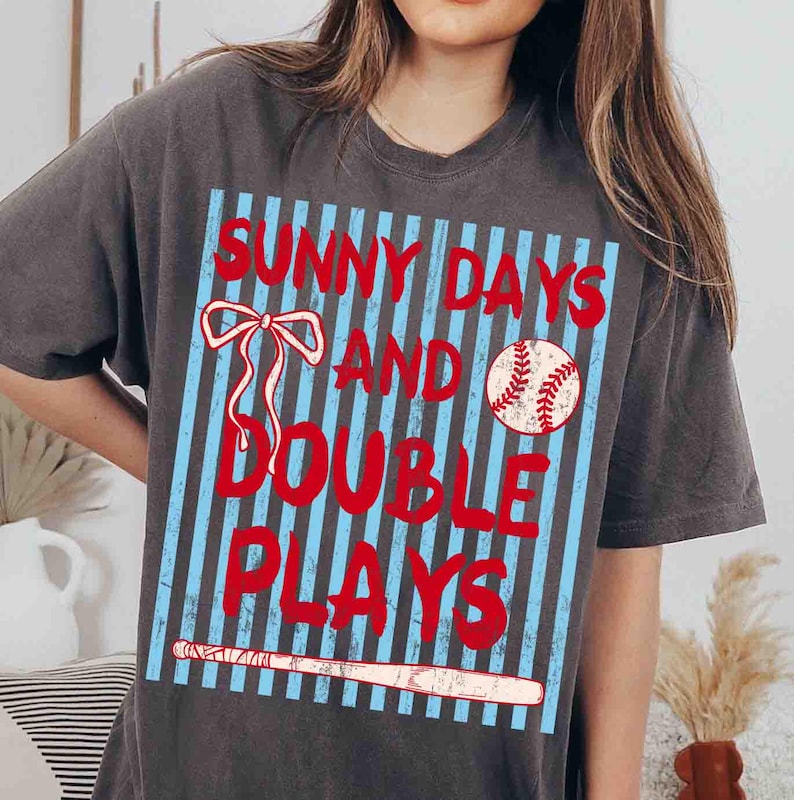 May include: Dark grey t-shirt with a baseball-themed graphic. The design features the text "SUNNY DAYS AND DOUBLE PLAYS" in red, along with a baseball, a bow, and a baseball bat, all on a blue striped background.