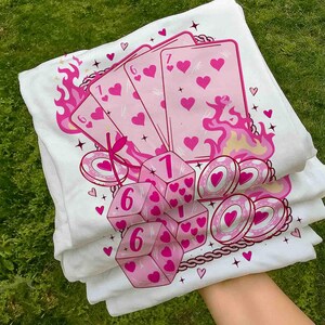 May include: White t-shirts with a pink graphic design featuring playing cards, dice, and poker chips. The design includes hearts, flames, and the numbers 1, 6, and 7. The shirts are stacked on top of each other.