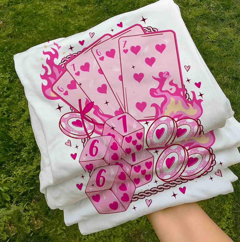 May include: White t-shirts with a pink graphic design featuring playing cards, dice, and poker chips. The design includes hearts, flames, and the numbers 1, 6, and 7. The shirts are stacked on top of each other.