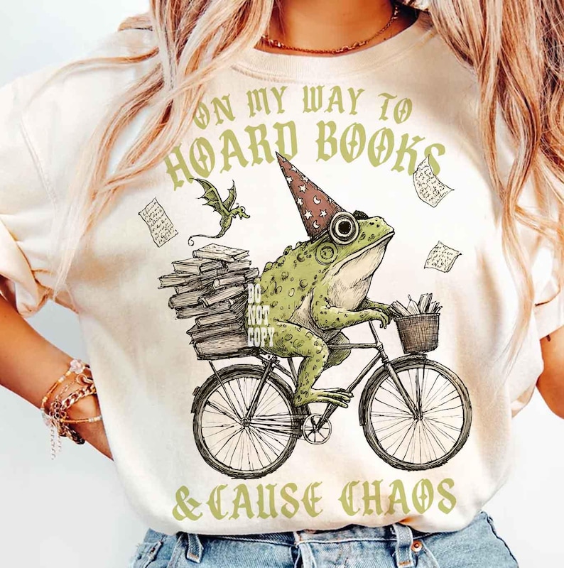 May include: Cream-coloured t-shirt with a whimsical illustration of a green frog riding a bicycle and wearing a wizard hat. The frog carries books and a basket. Text reads "ON MY WAY TO HOARD BOOKS & CAUSE CHAOS".