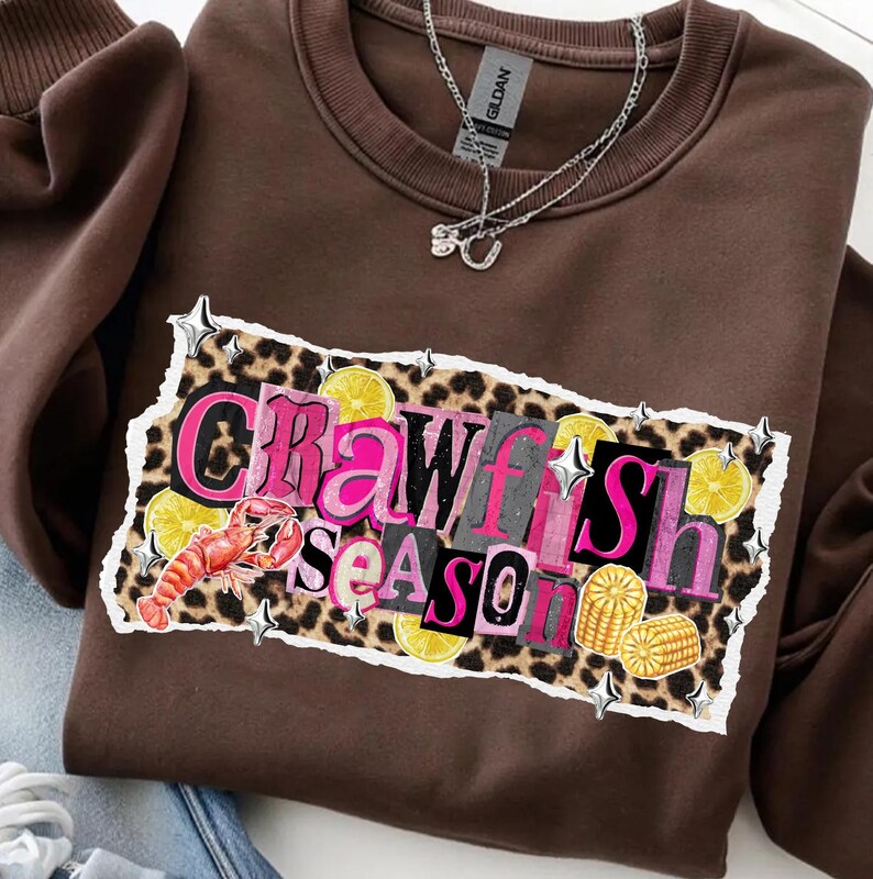 May include: A brown sweatshirt with the words "Crawfish Season" in a colorful, layered design. The design includes a crawfish, lemons, and corn. A silver necklace hangs on the front of the sweatshirt.