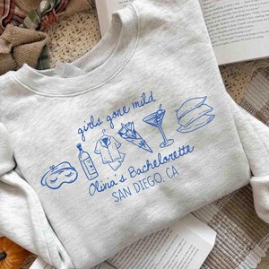 May include: A light gray sweatshirt with blue graphic text and illustrations. The text reads "girls gone mild" and "Olivia's Bachelorette SAN DIEGO, CA." Illustrations include a sleep mask, wine bottle, pajamas, flowers, a cocktail, and pillows.