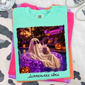 May include: A light teal Comfort Colors t-shirt featuring a graphic of a ghost in sunglasses relaxing in a pool with a drink. The graphic includes the text "Do Not Disturb" and "Summerween vibes". The shirt is stacked with other colorful shirts.