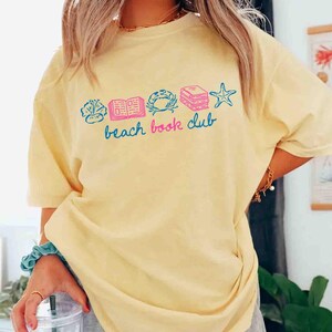 May include: A light yellow t-shirt with the words "beach book club" in blue script. The shirt features colorful illustrations of a seashell, book, crab, stack of books, and starfish.