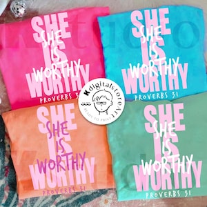 May include: Four colorful t-shirts: pink, blue, orange, and green. Each shirt features the text "SHE IS WORTHY" in white and pink, with "PROVERBS 31" below. A circular logo with a cartoon face is visible.