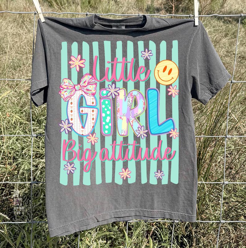 May include: A charcoal grey t-shirt featuring a graphic print with vertical mint green stripes. The design includes the text Little Girl Big attitude in pink, a large patterned bow, small flowers, and a yellow smiley face icon on the front.