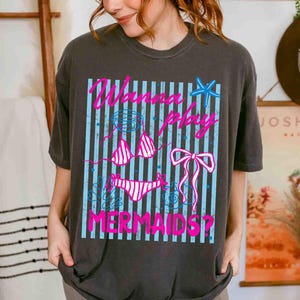 May include: Dark gray t-shirt featuring a graphic design with the text "Wanna Play Mermaids?" in pink. The design includes a bikini, bow, and starfish on a blue striped background. A casual, beach-themed tee.