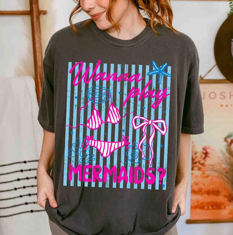May include: Dark gray t-shirt featuring a graphic design with the text "Wanna Play Mermaids?" in pink. The design includes a bikini, bow, and starfish on a blue striped background. A casual, beach-themed tee.