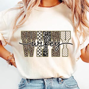 May include: A cream-coloured t-shirt featuring the word "Wifey" in a decorative design. The letters are filled with leopard print, black and white patterns, and a checkered pattern. The word "wifey" is written in a cursive font.