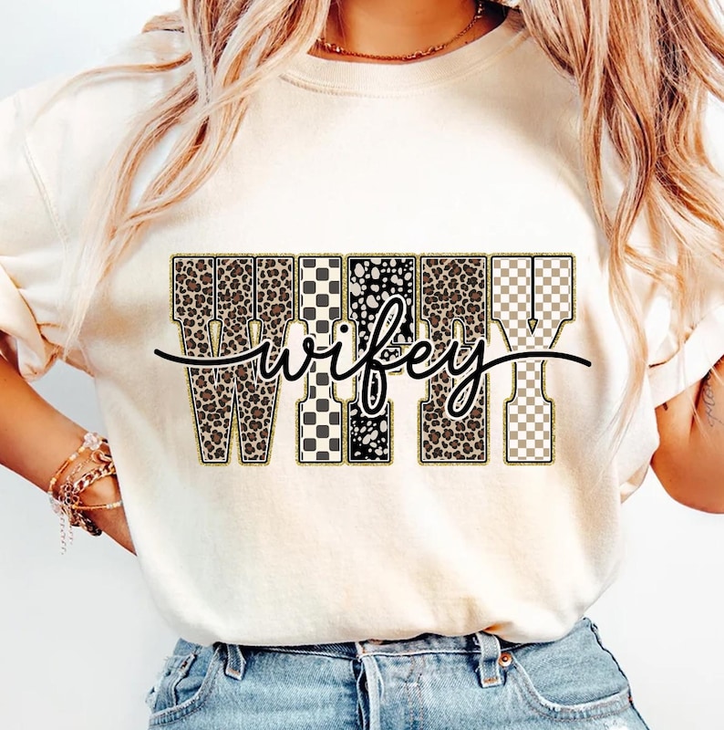 May include: A cream-coloured t-shirt featuring the word "Wifey" in a decorative design. The letters are filled with leopard print, black and white patterns, and a checkered pattern. The word "wifey" is written in a cursive font.