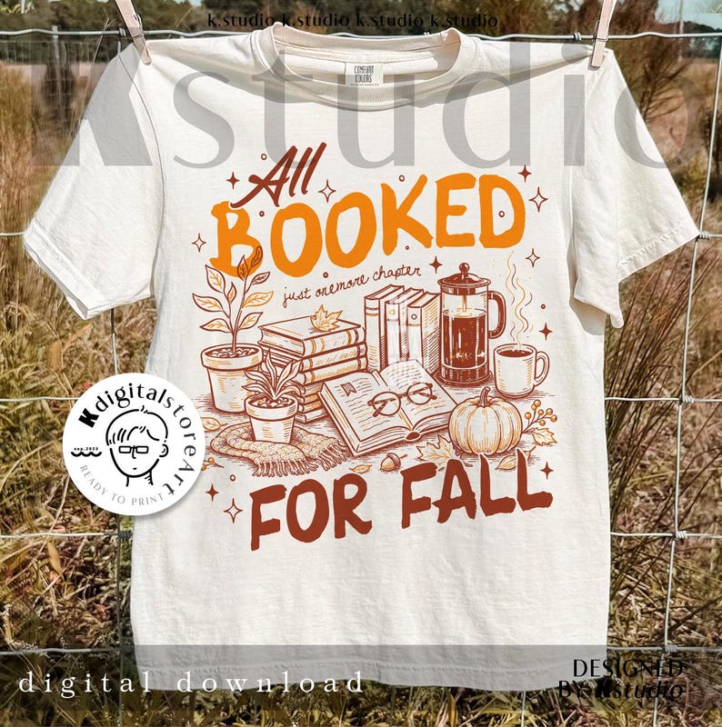 May include: Cream-colored t-shirt with the text "All Booked For Fall" in orange and brown. The design includes illustrations of books, a coffee pot, a pumpkin, and plants, evoking an autumn theme. The shirt is hanging on a clothesline.