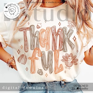K&ouml;nnte beinhalten: Ein cremefarbenes T-Shirt mit einem Grafikdesign, das das Wort Thankful in einer braun-orangen, krakeligen Schrift zeigt. Das Design enth&auml;lt Illustrationen von einem Truthahn, K&uuml;rbissen, Eicheln, Bl&auml;ttern und kleinen Rauten um den Text herum.