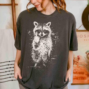 May include: Dark gray t-shirt featuring a white graphic of a raccoon giving the middle finger. The raccoon illustration is detailed with a distressed, textured effect. The shirt has a crew neck and short sleeves.