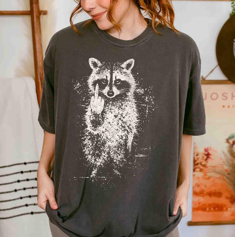 May include: Dark gray t-shirt featuring a white graphic of a raccoon giving the middle finger. The raccoon illustration is detailed with a distressed, textured effect. The shirt has a crew neck and short sleeves.