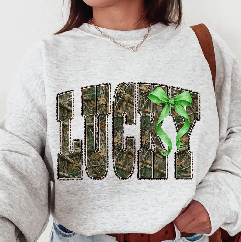 May include: A light gray sweatshirt with the word "LUCKY" in a camouflage pattern with a leopard print border. A green ribbon is attached to the letter "Y". The sweatshirt is a casual style.