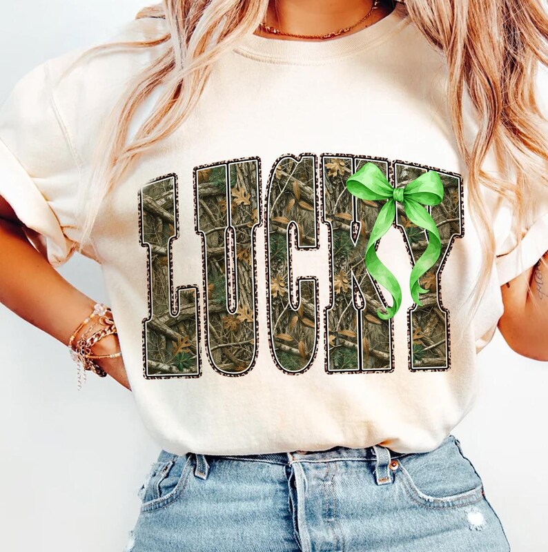 May include: Cream-colored sweatshirt with the word "LUCKY" in a camouflage pattern with a green bow. The letters are outlined in black. The sweatshirt is paired with blue jeans.