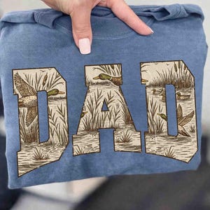 May include: A blue sweatshirt with the word "DAD" printed across the front. The letters are filled with a nature scene of ducks, reeds, and water. The sweatshirt is folded and held by a person.