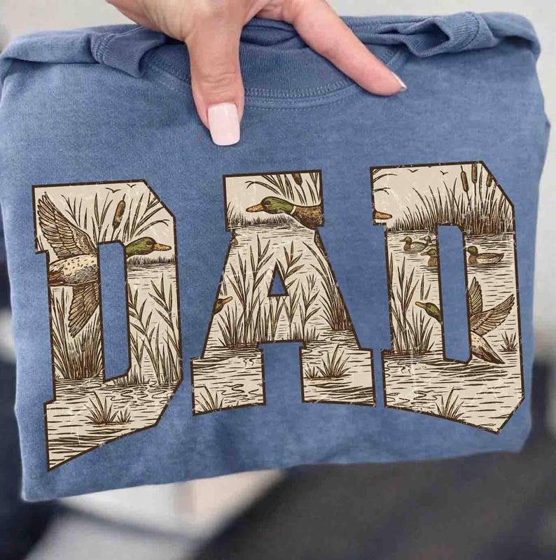 May include: A blue sweatshirt with the word "DAD" printed across the front. The letters are filled with a nature scene of ducks, reeds, and water. The sweatshirt is folded and held by a person.
