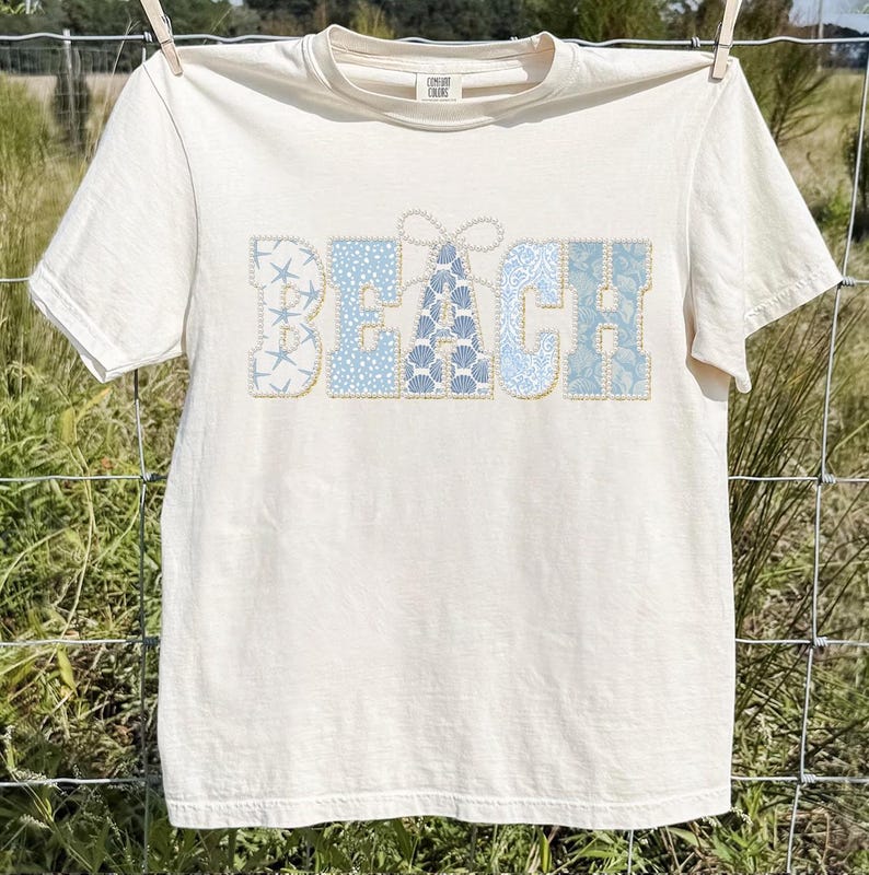 May include: A cream-coloured t-shirt displaying the word "BEACH" in light blue patterns of seashells and starfish. The t-shirt is hanging on a clothesline, suggesting a casual, beach-themed style.
