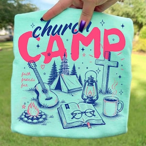K&ouml;nnte beinhalten: Hellblaues T-Shirt mit einem Grafikdesign mit den Worten "church camp" in Pink. Das Design umfasst eine Gitarre, ein Zelt, ein Kreuz, eine Laterne, eine Kaffeetasse und ein offenes Buch mit Brille. Die Worte "faith, friends, fun" sind ebenfalls enthalten.
