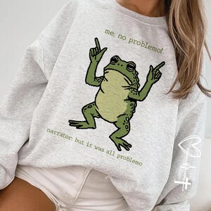 May include: A light gray sweatshirt featuring a cartoon frog with its arms raised. The frog is green with black spots. Text above the frog reads "me: no problemo!" and text below reads "narrator: but it was all problemo".