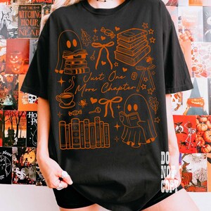 May include: Black t-shirt with an orange graphic featuring ghosts, books, bows, and the text "Just One More Chapter." The design includes a coffee cup, stars, and other Halloween-themed elements. The shirt is a casual, oversized fit.