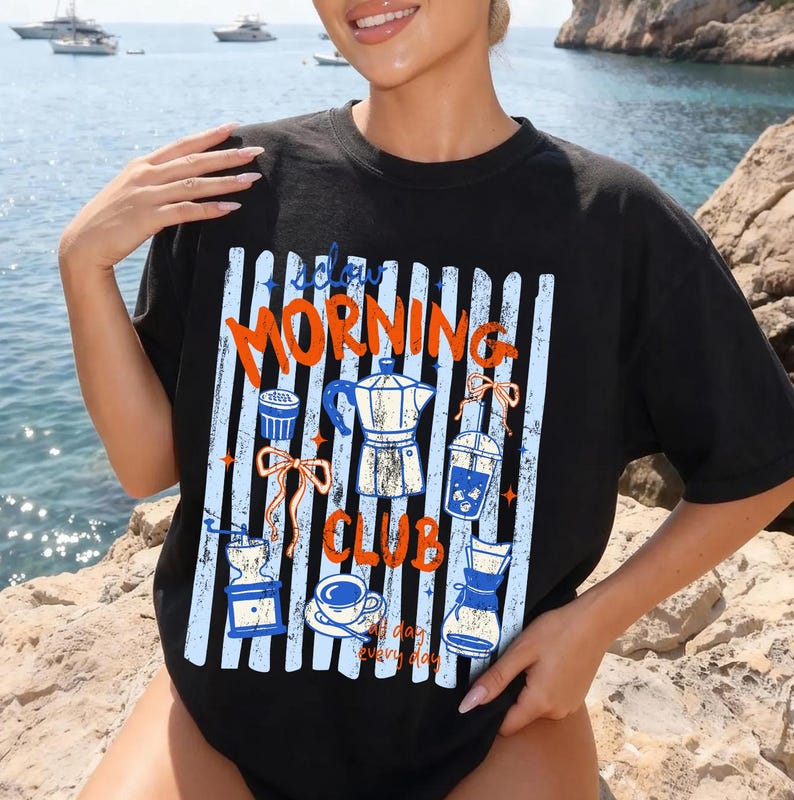 May include: Black t-shirt with a coffee-themed graphic. The design features the words "Slow Morning Club" in orange and white, along with illustrations of coffee-making equipment. The background has blue and white vertical stripes.