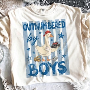 May include: Cream-colored t-shirt with a graphic featuring a goose wearing a red cap, holding a baseball and a monster truck. The text "OUTNUMBERED by BOYS" is in blue, with blue stars and stripes.