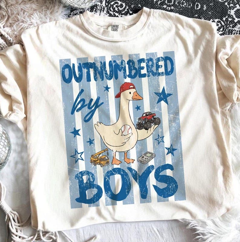 May include: Cream-colored t-shirt with a graphic featuring a goose wearing a red cap, holding a baseball and a monster truck. The text "OUTNUMBERED by BOYS" is in blue, with blue stars and stripes.