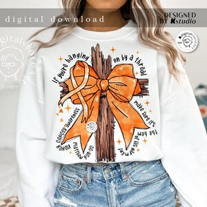 May include: White sweatshirt with an orange ribbon tied around a brown wooden cross. The text "If you're hanging on by a thread make sure it's the hem of His garment" is printed on the sweatshirt.  Cancer awareness, Matthew 9:20-22, and KIDNEY are also printed on the sweatshirt.