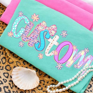 May include: A turquoise t-shirt with the word "Custom" in a colorful, floral design. The word is outlined in various colors, with polka dots, leopard print, and a bow. The shirt is folded on top of pink fabric.
