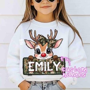 May include: White sweatshirt featuring a cartoon reindeer with a camouflage bow and a red nose. The reindeer holds a sign with the name "EMILY" in white letters. The sweatshirt is for children.